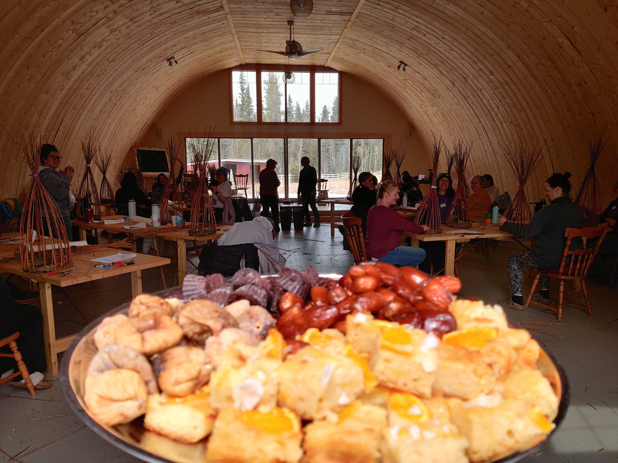 A platter of Roni's food in the foreground, weavers at their willow baskets, and the forest through the lodge windows