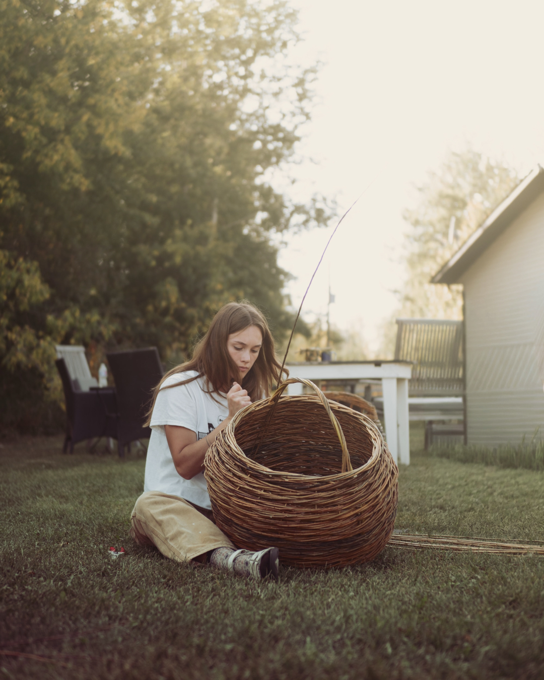 A weaver working the willow spokes of a large basket taking shape on the lawn