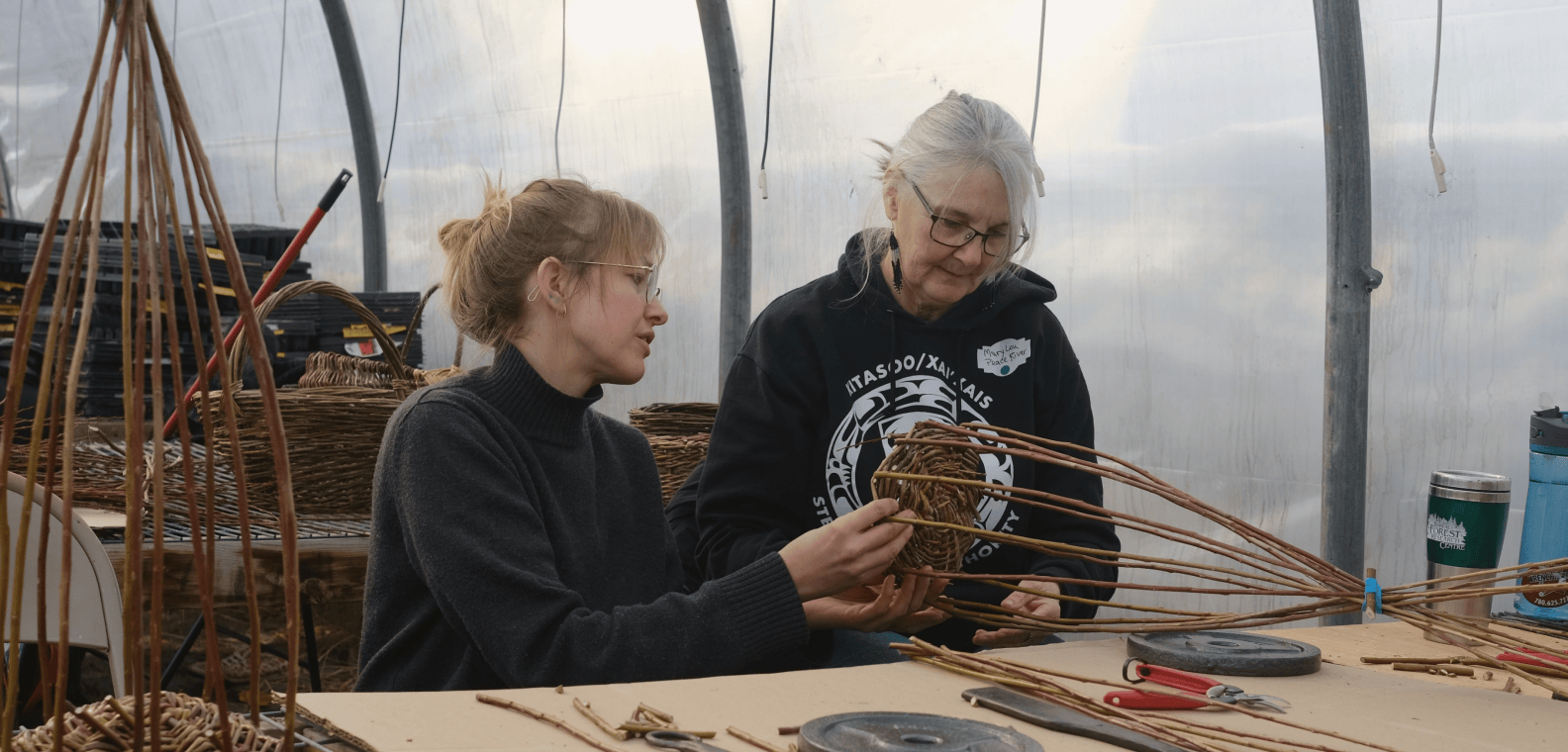 Two weavers at a long table in a greenhouse, working together on a willow basket
