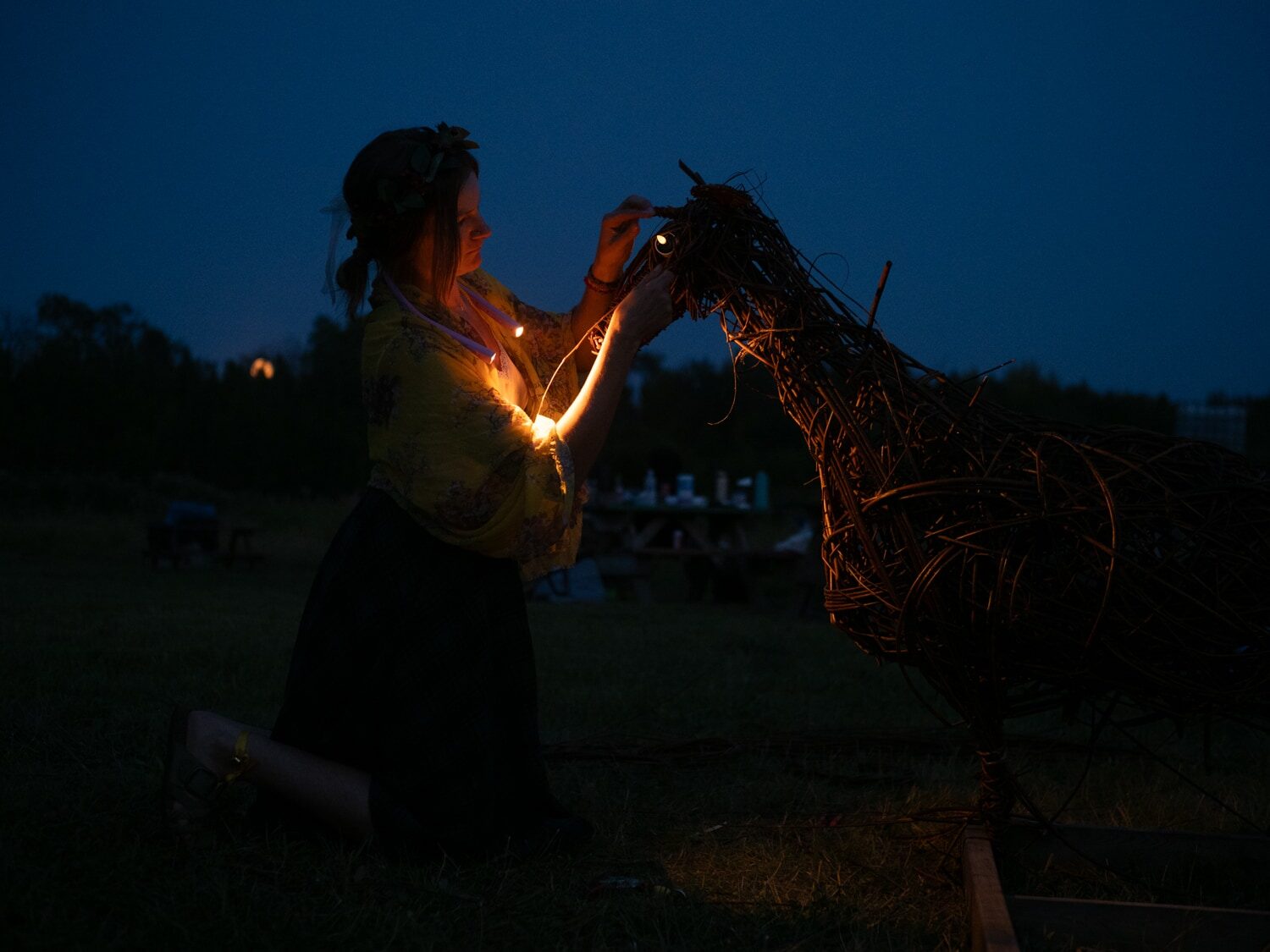 The willow goat at dusk with glowing LED candle eyes, a weaver beside it